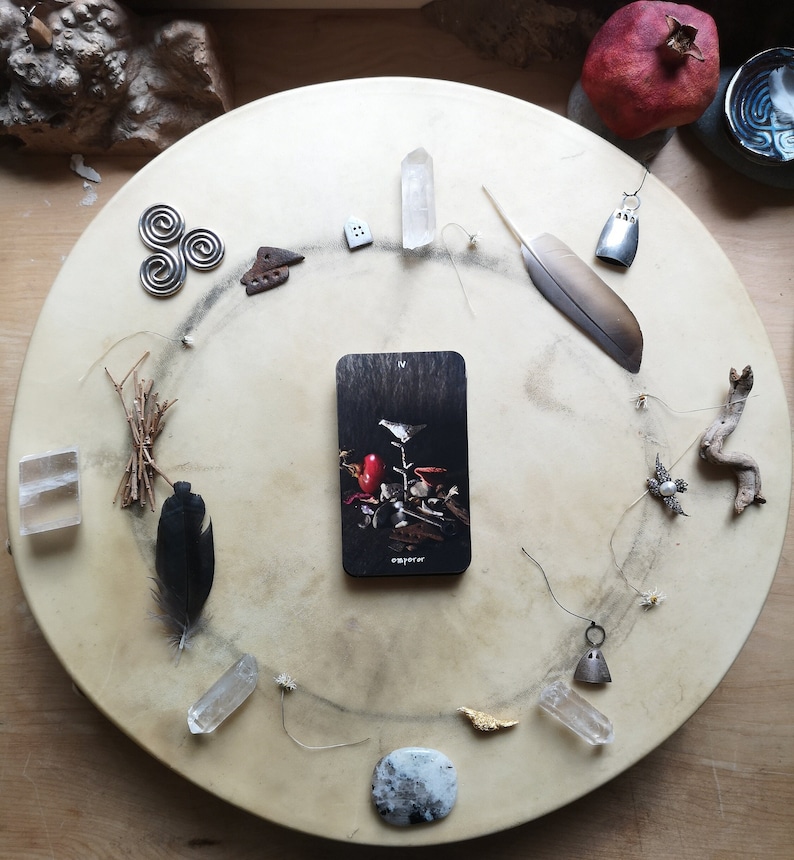 Pu&ograve; includere: Una disposizione circolare di oggetti spirituali su una superficie marrone chiaro. Include cristalli, piume, una carta dei tarocchi, piccoli ciondoli di metallo e un melograno. La carta dei tarocchi presenta un uccello e la parola "emperer".