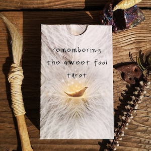 May include: A white card with the text "remembering the sweet fool tarot" in black script. The card features a white, feathery background with a golden bird in the center.