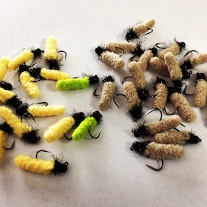 May include: A selection of artificial fishing flies. The flies are in shades of yellow, green, and beige, with black detailing and curved hooks. These flies are designed to imitate insects.