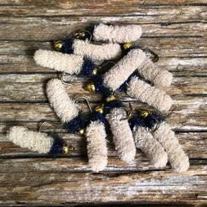 May include: A selection of handmade fishing flies. Each fly has a beige, fuzzy body, a dark blue tail, a gold-coloured bead, and a curved hook. The flies are arranged on a rustic, wooden surface.