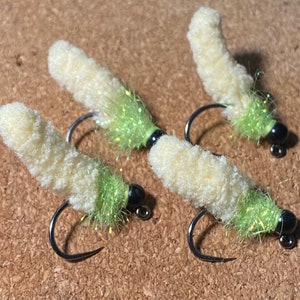 Może przedstawiać: Cztery ręcznie robione muchy wędkarskie. Każda mucha ma kremowy, puszysty korpus, jasnozielony ogon i czarną główkę z koralików. Muchy są przymocowane do srebrnych haczyków, przeznaczonych do łowienia ryb.