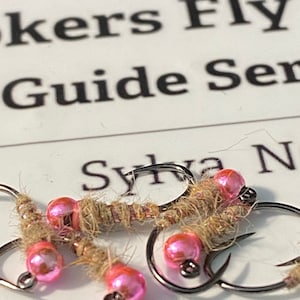 Può includere: Primo piano di diverse mosche da pesca con perline rosa iridescenti e corpi in filo marrone. Gli ami sono argentati e le mosche sono disposte su una superficie bianca con la scritta "Sylva N".