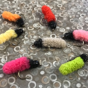 Może przedstawiać: Kolekcja kolorowych much wędkarskich. Każda mucha ma puszysty, cylindryczny korpus w odcieniach pomarańczu, żółci, czerwieni, różu, beżu i zieleni, z czarną główką i zakrzywionym haczykiem. Muchy są przeznaczone do wędkowania.