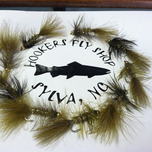 May include: Fishing flies with olive green and black feathers, arranged around a circular logo. The logo features a black fish silhouette and reads "Hookers Fly Shop, Sylva, NC". The flies have gold-colored beads.