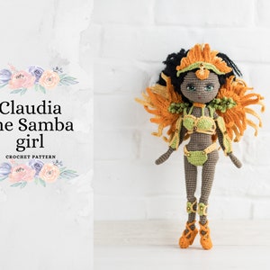 May include: Crocheted doll with brown skin, green and gold costume, and orange feathers. The doll is wearing a headdress with orange feathers and a green and gold costume. The doll is standing on a white surface. The text "Claudia the Samba girl" is written in black on a white background with pink and purple flowers.