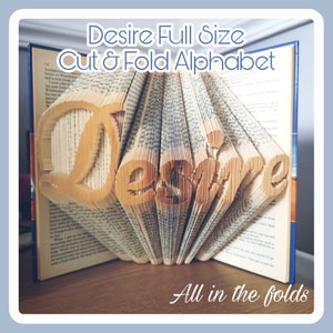 Può includere: Un libro con la parola "Desire" piegata nelle pagine. Il libro è aperto e le pagine sono piegate per creare la parola. Il titolo dell'immagine è "Desire Full Size Cut & Fold Alphabet" e il testo "All in the folds" è in basso nell'immagine.