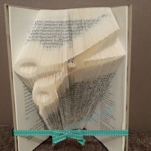 May include: A book folded into the shape of a bird, with a teal ribbon tied around it. The book is standing upright on a brown surface. The word "Pattern" is written in blue above the book.