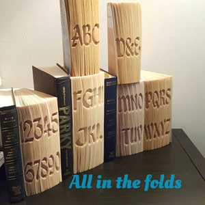 May include: A set of seven books with the alphabet cut and folded into the pages. The books are arranged in a row on a wooden surface. The text on the books reads "Cut and fold Mini P" and "All in the folds".