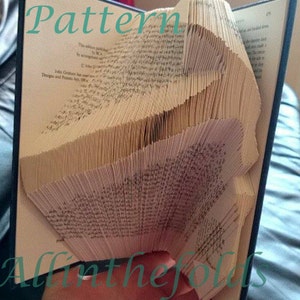 May include: A book with pages folded into a 3D pattern, creating a unique and intricate design. The book is open and the folded pages are visible. The text "Pattern" is written in green on the front cover of the book. The text "All in the folds" is written in green at the bottom of the image.