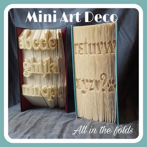 May include: Two books with the alphabet folded into the pages. The book on the left has the letters a-q and the book on the right has the letters r-z. The title "Mini Art Deco" is at the top of the image and the text "All in the folds" is at the bottom.