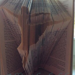 Pode incluir: Escultura de livro dobrado com a palavra "Pattern" em escrita turquesa no topo e "Allinthefolds" na parte inferior. As páginas são dobradas para criar um design tridimensional, mostrando a textura do papel.