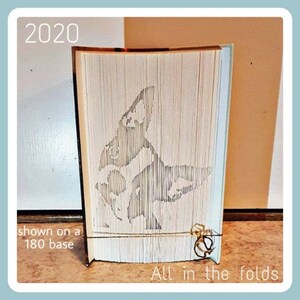 May include: A white book with a folded design of a dog, shown on a 180 base. The book is titled "All in the folds" and the year 2020 is printed on the top left corner.