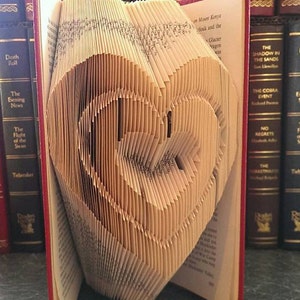 May include: A book folded into the shape of a heart. The book is open and the pages are folded to create a three-dimensional heart shape. The book is sitting on a shelf with other books.