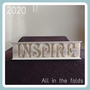 以下が含まれることがあります： 「INSPIRE」という言葉の形に折り畳まれた白い本。本は濃い茶色の表面の上に置かれています。画像の左上隅には2020年と書かれています。本の真下には「All in the folds」というテキストが書かれています。