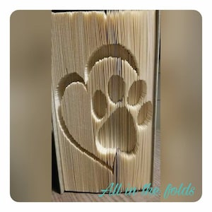 Peut inclure: Un livre avec un cœur et une empreinte de patte pliés dans les pages. Le livre est beige et le motif est dans une nuance de beige plus claire. Le texte "All in the folds" est visible en bas de l'image.