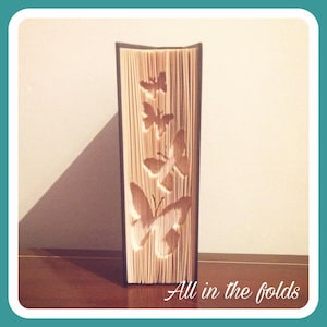 Op de afbeelding: Een decoratief boek met vlinderuitsparingen. De pagina's zijn gevouwen om de vlindervormen te creëren. Het boek staat rechtop op een houten oppervlak. De tekst "All in the folds" staat onderaan.