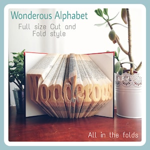 May include: A book art piece with the word "Wondrous" folded into the pages. The book is open and the pages are fanned out to create a 3D effect. The book is on a wooden table with a green plant in a white pot in the background.