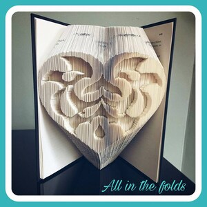 May include: A book with pages folded into a heart shape. The heart is made of intricate patterns and designs. The book is open and the pages are folded to create a three-dimensional heart. The text "All in the folds" is visible at the bottom of the image.