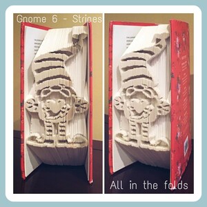 May include: A red book with a white gnome folded into the pages. The gnome has a striped hat and is standing with its arms outstretched. The text "Gnome 6 - Stripes" is at the top of the image and "All in the folds" is at the bottom.