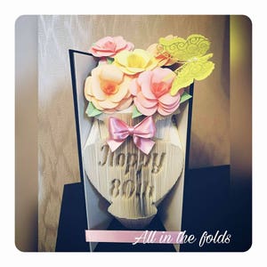 Könnte beinhalten: Eine Papierbastelvase mit einer rosa Schleife und Blumen in Rosa-, Gelb- und Pfirsichtönen. Die Vase ist mit den Worten "Happy 80th" verziert.