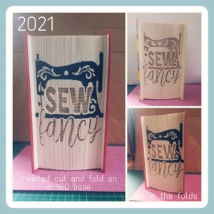 May include: A red book with a black sewing machine and the words "Sew Fancy" cut out of the pages. The book is folded into a 3D sculpture.