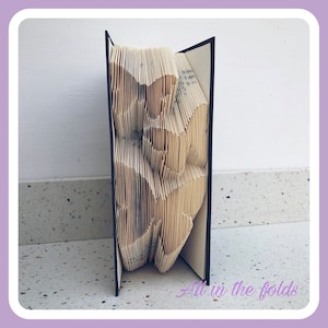 Puede incluir: Un libro doblado en una escultura tridimensional de una flor. El libro está abierto y las páginas están dobladas para crear la forma de una flor. El libro está de pie sobre una mesa.