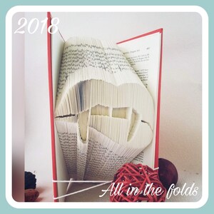 Może przedstawiać: Książka ze stronami złożonymi w kształt ust z kłami. Książka jest czerwona i biała i ma napis "All in the folds" wydrukowany na dole. Rok 2018 jest napisany na górze po lewej stronie obrazu.
