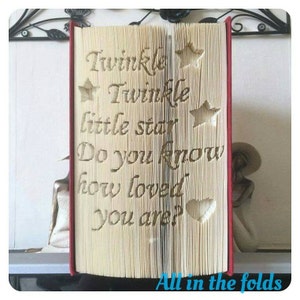 Op de afbeelding: Een boek met de woorden "Twinkle Twinkle little star Do you know how loved you are?" gevouwen in de pagina's. Het boek is rood en heeft een hart en sterren die uit de pagina's zijn geknipt.
