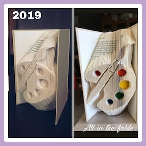 Peut inclure: Une sculpture d'art de livre représentant une palette de peinture et un pinceau, pliés à partir des pages d'un livre. La sculpture est blanche avec des éclaboussures de peinture colorées sur la palette. La sculpture est intitulée "All in the folds".
