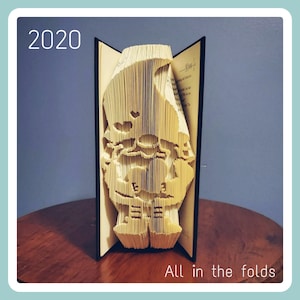 May include: A book with a folded paper design of a gnome, the book is open and the design is visible. The book is on a wooden surface. The text "2020" is visible in the top left corner of the image. The text "All in the folds" is visible in the bottom right corner of the image.