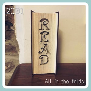 May include: A book with the word "READ" folded into the pages. The book is standing upright on a wooden surface. The text "2020" is in the top left corner of the image. The text "All in the folds" is at the bottom of the image.