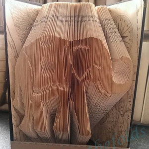 Pode incluir: Um livro dobrado na forma de um elefante. O livro é feito de páginas antigas e amareladas e está em pé sobre uma superfície escura. O livro é intitulado "Pattern" e está rodeado por outros livros.