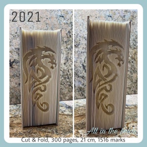 May include: A beige paper book folded into a 3D dragon sculpture. The book is 21 cm tall and has 300 pages. The text "2021" and "Cut & Fold, 300 pages, 21 cm, 1516 marks" are visible on the image.