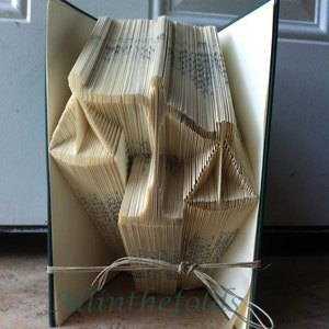 May include: A book folded into a three-dimensional shape, resembling a geometric design. The book is bound in green and has a cream-colored interior. The book is tied with a brown string.