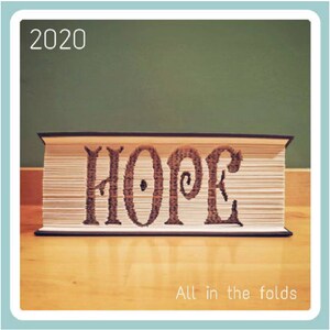 May include: A book folded into the shape of the word "HOPE" with the year 2020 above it. The book is sitting on a wooden surface. The text "All in the folds" is below the book.