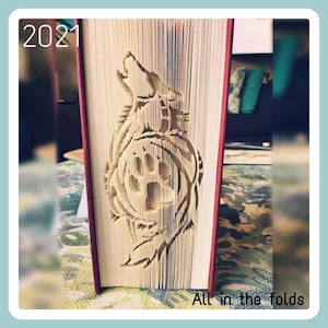 May include: A book with a wolf howling design cut out of the pages. The design is intricate and detailed, with the wolf's fur and the surrounding foliage all carefully crafted. The book is closed, and the design is visible from the side. The book is red and has the year 2021 printed on the spine. The text "All in the folds" is printed below the book.