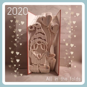 May include: A book folded into a 3D sculpture of a gnome holding a heart. The book is open and the gnome is standing in the middle of the pages. The background is a light blue with white hearts.