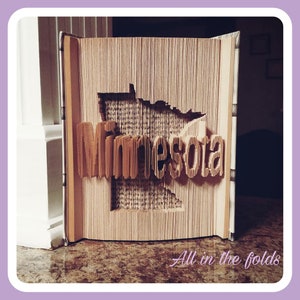 May include: A book art sculpture of the state of Minnesota with the word "Minnesota" folded into the book. The book is open and the sculpture is on a brown surface. The text "All in the folds" is written below the sculpture.