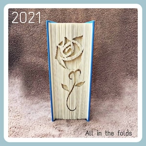 May include: A book with a blue spine has been folded into a 3D sculpture of a rose. The rose is cut out of the pages of the book, creating a delicate and intricate design. The book is standing upright on a brown surface. The text "All in the folds" is printed below the book.
