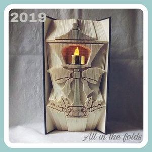 May include: A book folded into the shape of a lantern with two lit candles inside. The book is open and the pages are folded to create the shape of the lantern. The text "All in the folds" is visible at the bottom of the image. The year 2019 is visible in the top left corner of the image.