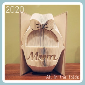 May include: A book folded into a three-dimensional sculpture of a basket with a bow on top. The basket says "Mom" with a heart in the middle. The sculpture is on a wooden surface. The text "All in the folds" is below the sculpture. The year 2020 is in the top left corner of the image.
