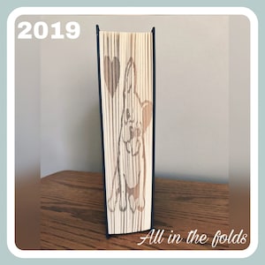 May include: A book art sculpture of a dog with a heart, folded from the pages of a book. The sculpture is standing on a wooden surface. The text "All in the folds" is visible at the bottom of the image.