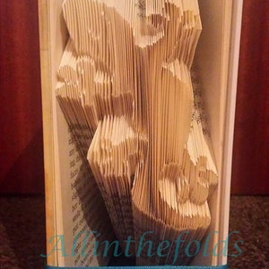May include: A book folded into a 3D sculpture of a bear. The book is bound in brown and white and has a blue ribbon tied around it. The title of the book is "The Children's Story" by  James Herriot. The sculpture is titled "All in the Folds".