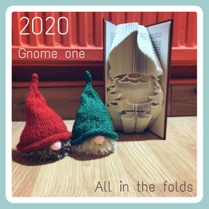 May include: Two knitted gnomes, one red and one green, are sitting in front of a book folded into the shape of a gnome. The book is open and the pages are folded into a 3D design. The text "2020 Gnome one All in the folds" is visible in the image.