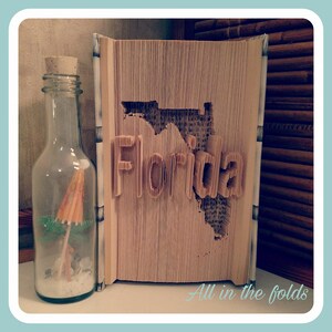 May include: A book folded into the shape of the state of Florida with the word "Florida" written in the folds. The book is standing on a table next to a clear glass bottle with a cork stopper and a small beach umbrella inside.
