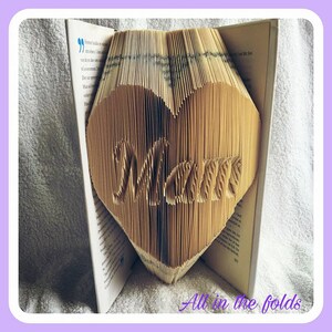 Könnte beinhalten: Ein herzförmiges Buchkunstwerk mit dem Wort "Mama" in die Seiten geschnitzt. Das Buch ist geöffnet, das Herzdesign steht aufrecht. Der Text "All in the folds" befindet sich am unteren Bildrand.