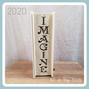 Könnte beinhalten: Ein Buch mit dem Wort "IMAGINE" in den Rücken geschnitten. Das Buch steht aufrecht auf einer Holzoberfläche. Das Jahr 2020 ist in der oberen linken Ecke des Bildes geschrieben. Der Text "All in the folds" steht unter dem Buch.