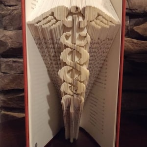 May include: A book with pages folded into a three-dimensional sculpture of the medical symbol, the Rod of Asclepius. The book is open and the sculpture is visible from the front. The book is on a wooden surface. The text "All in the folds" is visible at the bottom of the image.