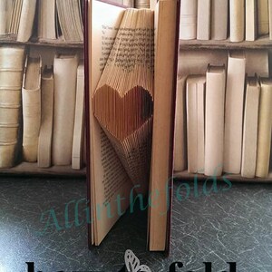 May include: A book with a heart shaped fold in the pages. The book is standing upright on a table with a bookshelf in the background. The text on the book reads "Video tutorial how to fold a basic heart".