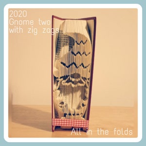 May include: A folded book art piece featuring a gnome design. The book is red with a gingham ribbon and the text "2020 Gnome two with zig zags" and "All in the folds". The gnome has a beard and hat.